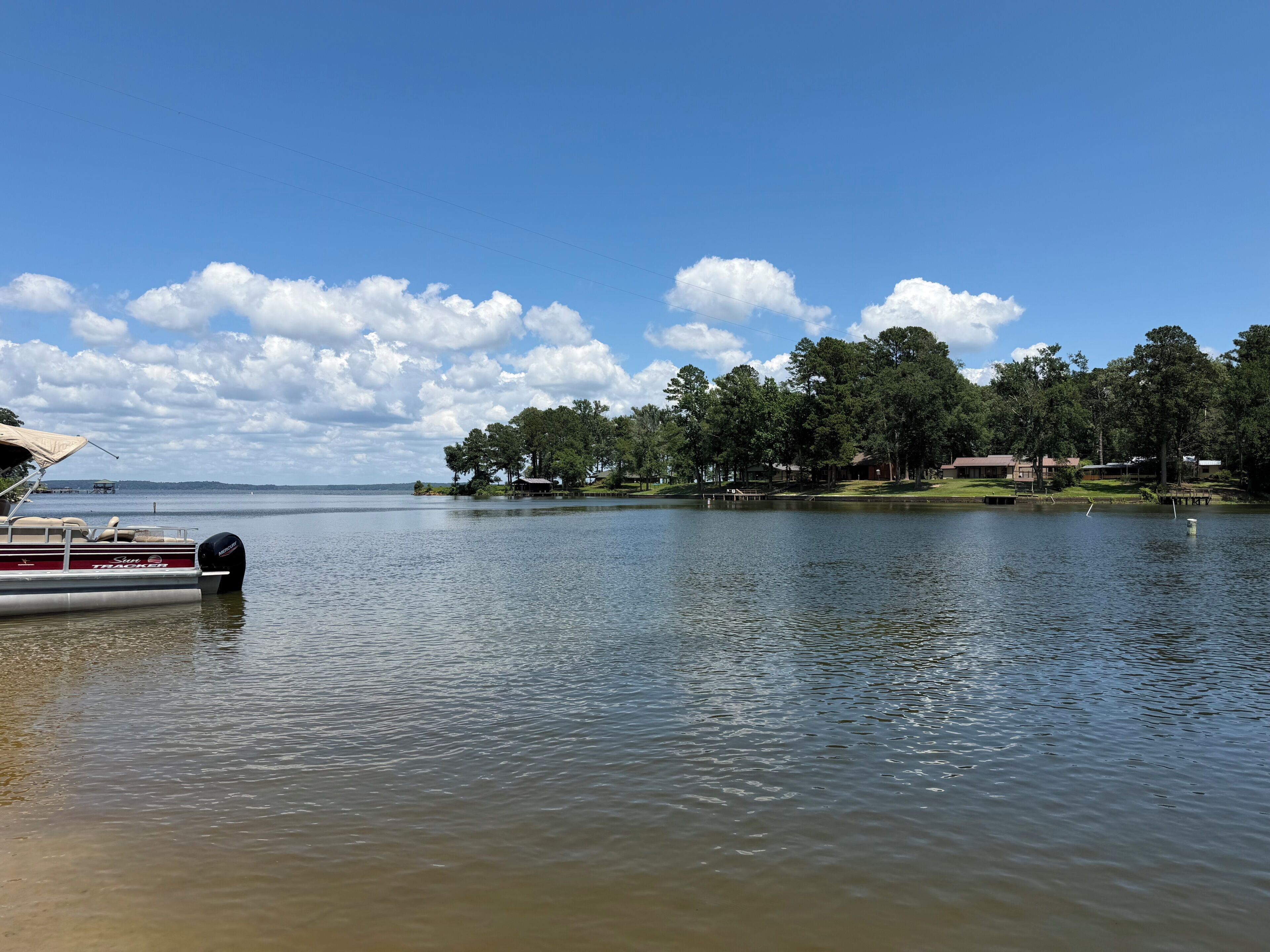Um lago com um barco atracado à esquerda, rodeado por árvores e alguns edifícios ao fundo.