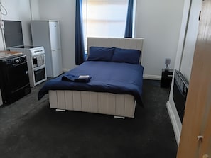 Ein Schlafzimmer mit einem Bett, einer kleinen Küchenzeile, einem Heizkörper und einem Fenster mit Vorhängen.