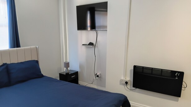 Un dormitorio con cama, mesita de noche, lámpara, calentador de pared y toma de corriente.