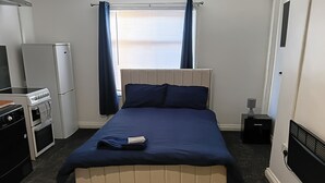 Un dormitorio con cama, una kitchenette pequeña, una ventana con cortinas y un radiador.