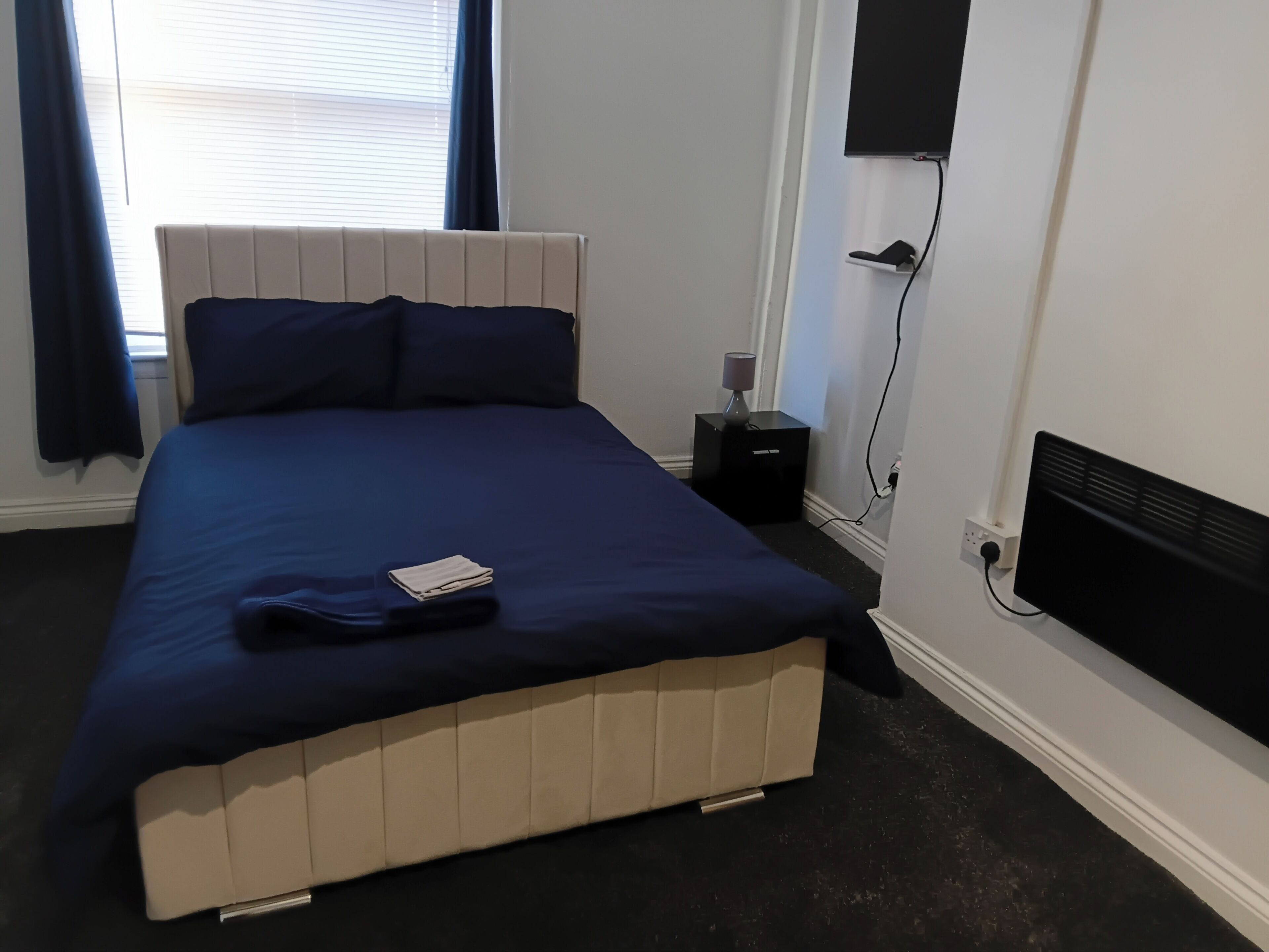 Un lit avec une tête de lit, une table de chevet avec une lampe, un téléviseur fixé au mur et une fenêtre avec des rideaux.