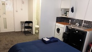Habitación de hotel compacta con cama, lavadora, lavamanos pequeño y una silla.