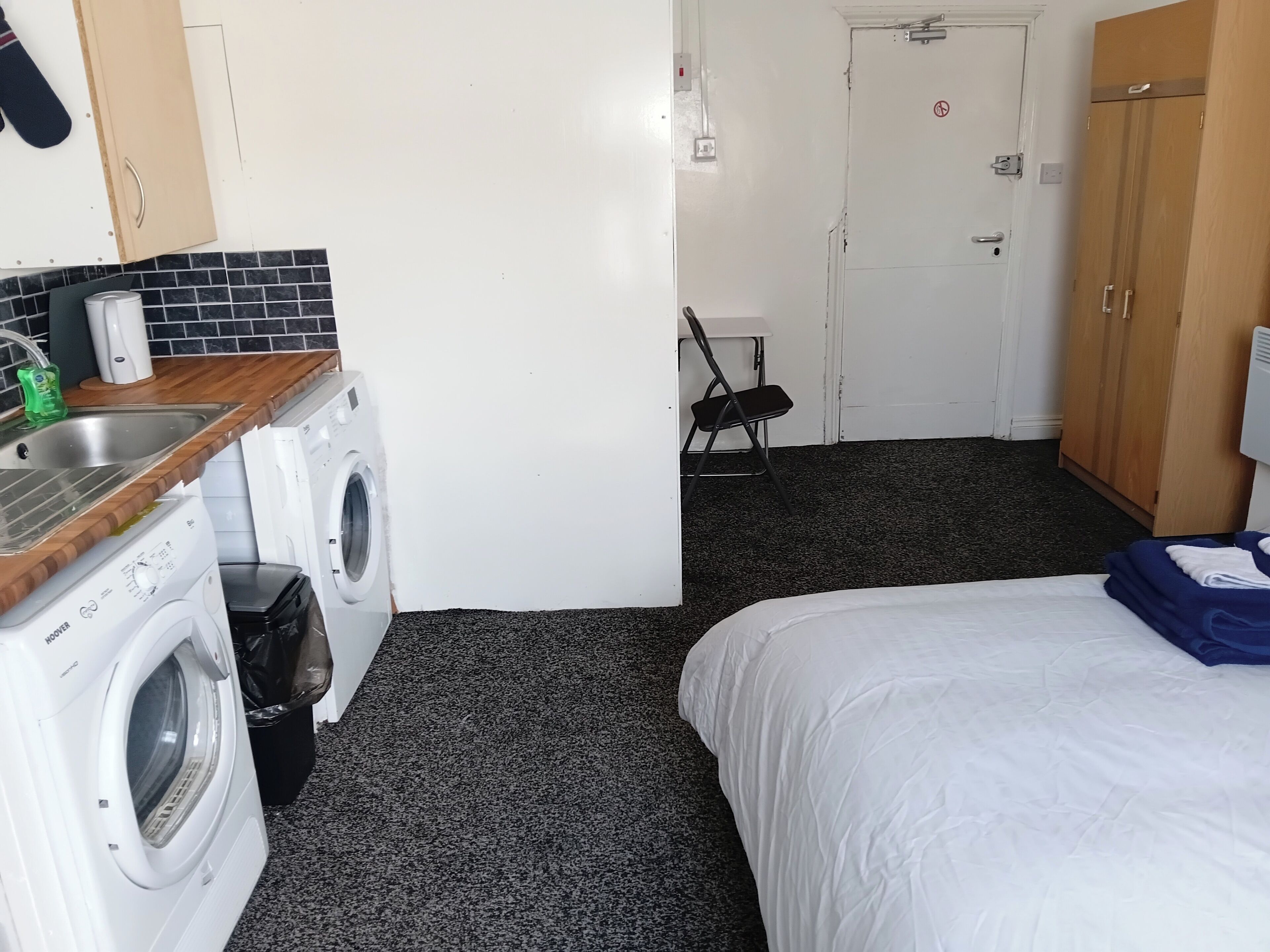 A compact hotel room with a washing machine, a bed, a chair, and a door.