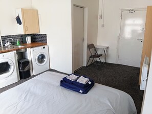 Hotellihuone, jossa on sänky, pesukone, pieni pöytä ja tuoli sekä ovi.