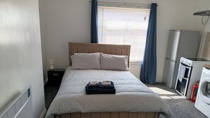 A neatly made bed with pillows, a bedside table with a lamp, a window with curtains, and a kitchenette area with a washing machine. - Studio Sleeps 2 With Parking (Oldbury)