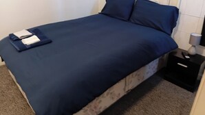A bed with a blue bedspread and pillows, a white headboard, a nightstand with a lamp, and a remote control. - Serene Studio Retreat in Oldbury (Oldbury)