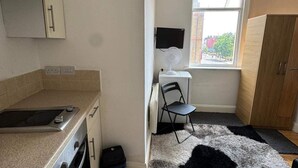 A compact kitchen with a microwave, oven, and a small countertop. There's a chair and a desk with a fan, a window with a view of the street, and a wardrobe. - Upper Holloway Studio, 1 Min to Station, Sleeps 2 (London)