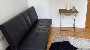 A black leather sofa, a small wooden table with a bottle and a roll of paper towels, and a grey and black shag rug on a wooden floor. - Upper Holloway Studio, 1 Min to Station, Sleeps 2 (London)