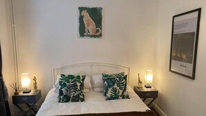Ett sovrum med en vit säng, gröna kuddar med ett bladmotiv, ett konstverk med leopardmönster, en inramad tavla och två sängbord med lampor.
