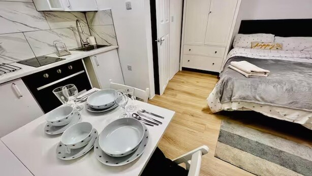 A compact room with a kitchenette, a dining table set with plates and cutlery, a bed with a grey duvet, and a striped rug. - Studio Sleeps 2 With Shared Hot Tub (London)