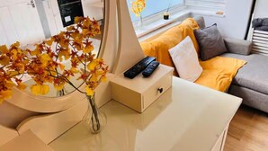 A modern living room with a white desk, a vase with yellow flowers, a remote control, and a grey sofa with a yellow throw blanket. - Studio Sleeps 2 With Shared Hot Tub (London)