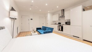 A modern hotel room with a large bed, a blue sofa, a small table, a lamp, a kitchenette, and a door.