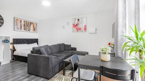 Uma sala de estar moderna com um sofá seccional cinza, uma pequena mesa de jantar com cadeiras, uma cama com cabeceira cinza e obras de arte nas paredes.
