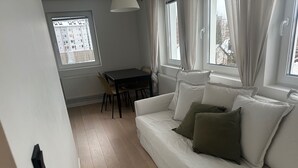 Ein Zimmer mit einer weißen Couch, einem Esstisch, Stühlen und großen Fenstern mit Vorhängen.