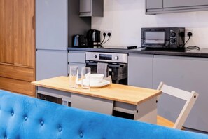 Eine Küchenzeile mit Mikrowelle, Kaffeemaschine und Backofen. Ein Tisch mit Gläsern und einem Teller steht bereit. Eine blaue, gepolsterte Bank befindet sich im Vordergrund.
