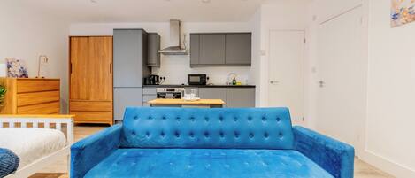 A modern living room with a blue sofa, a wooden coffee table, a remote control, a kitchen area with cabinets and appliances, and a white door.