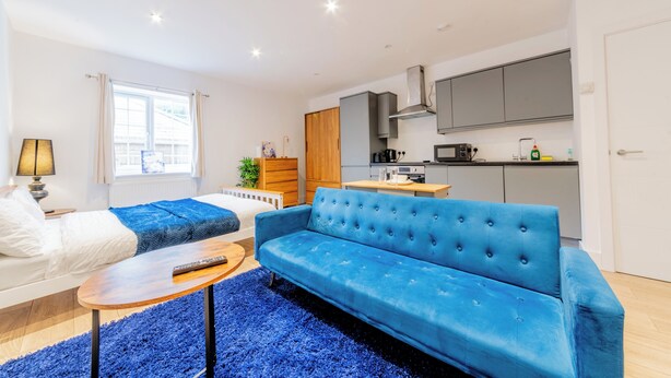 A modern living room with a blue sofa, a wooden coffee table, and a kitchen area with grey cabinets and a microwave. - Sleeps 2 - Wifi - Paid Parking - Central Studio (Maidstone)
