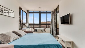 Una habitación de hotel con una cama, un escritorio, una silla, un televisor y ventanas amplias.