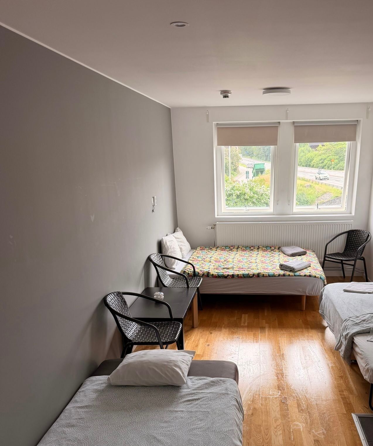 A small, well-lit room with two single beds, a table with chairs, and a window with a view of greenery and a road.