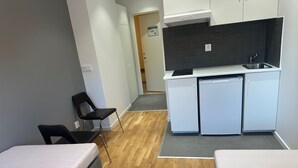 Una habitación compacta con dos camas, kitchenette, una silla negra y piso de madera.