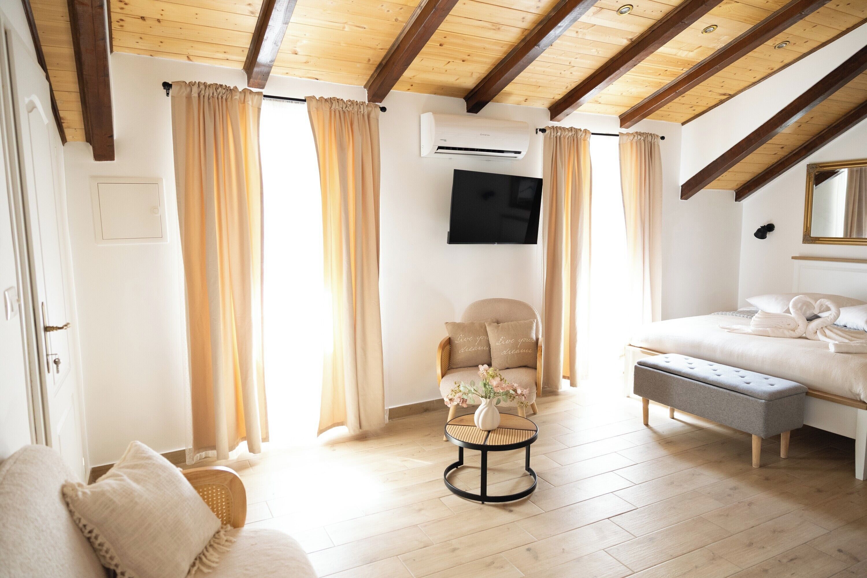 A room with a bed, a chair, a small round table with a vase, and a television on the wall.