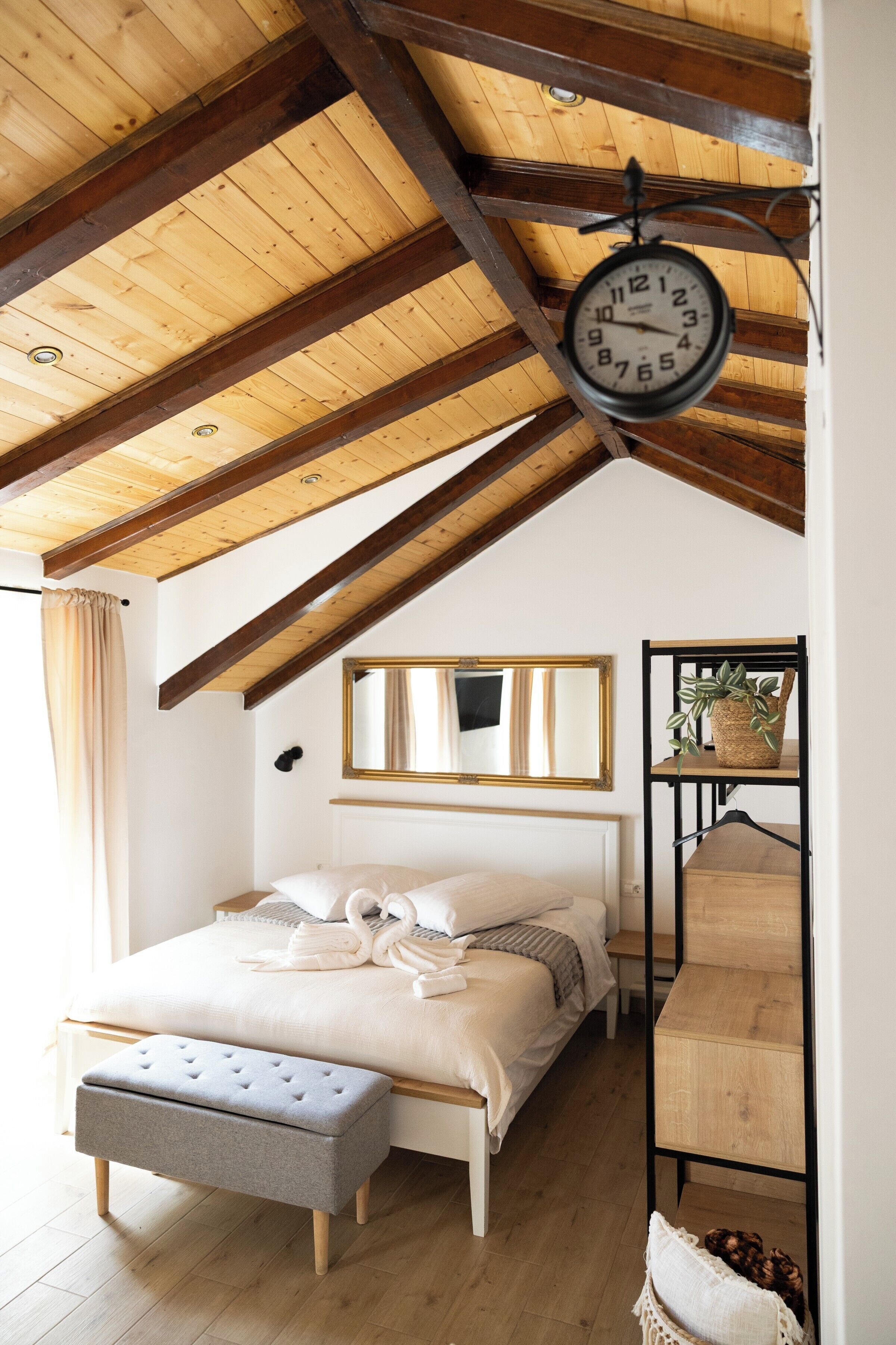 Ein Schlafzimmer mit Holzboden, einem Bett mit grauer Sitzbank, einer Wanduhr und einem Regal mit einer Pflanze.