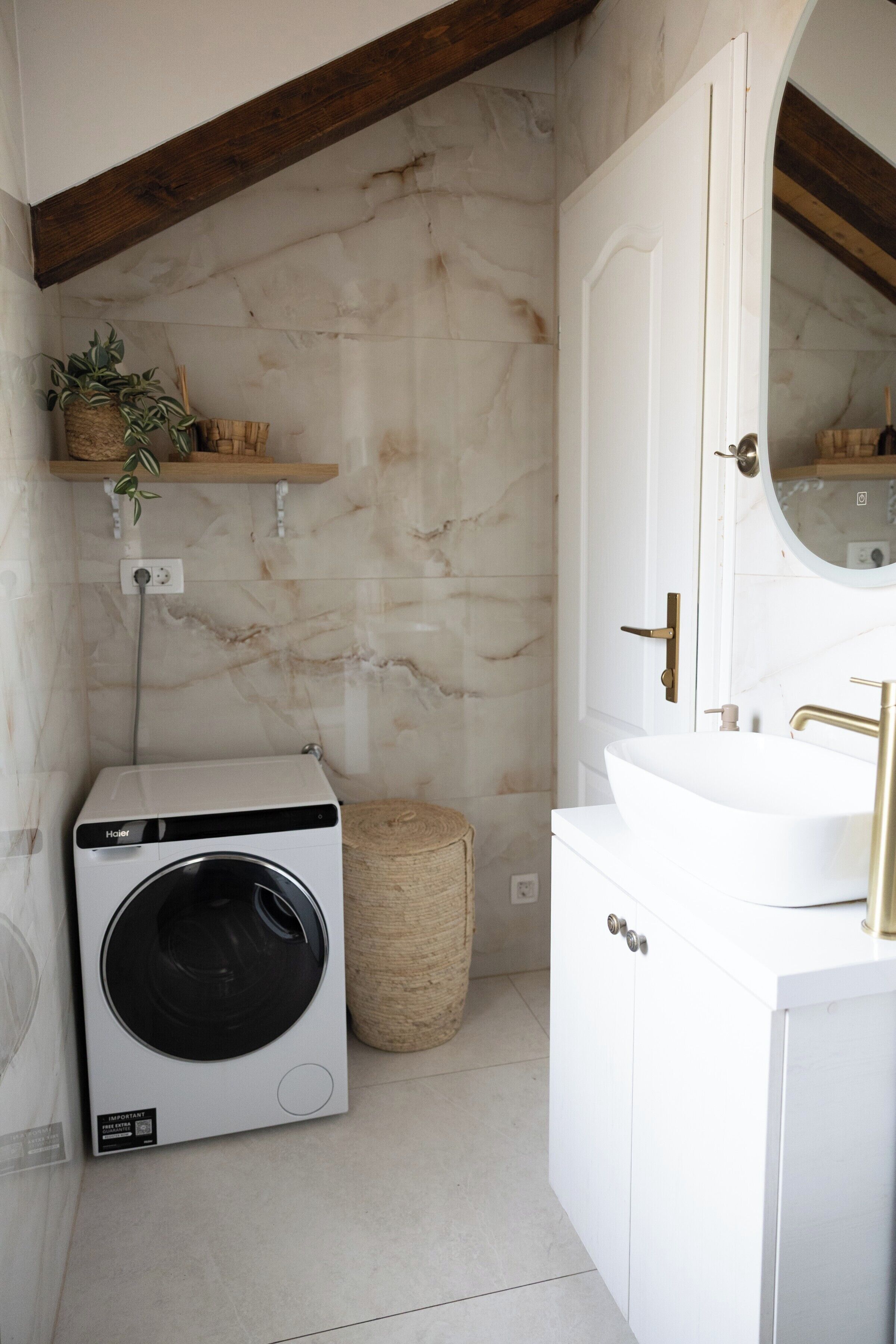 A bathroom with a white washing machine, a wooden shelf with plants, a round woven basket, and a white vanity with a gold faucet.