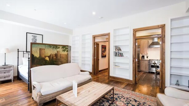 Una sala moderna con un sofá blanco, una mesa de centro y un cuadro en la pared. Se observan estantes empotrados y una cocina a través de una puerta abierta.