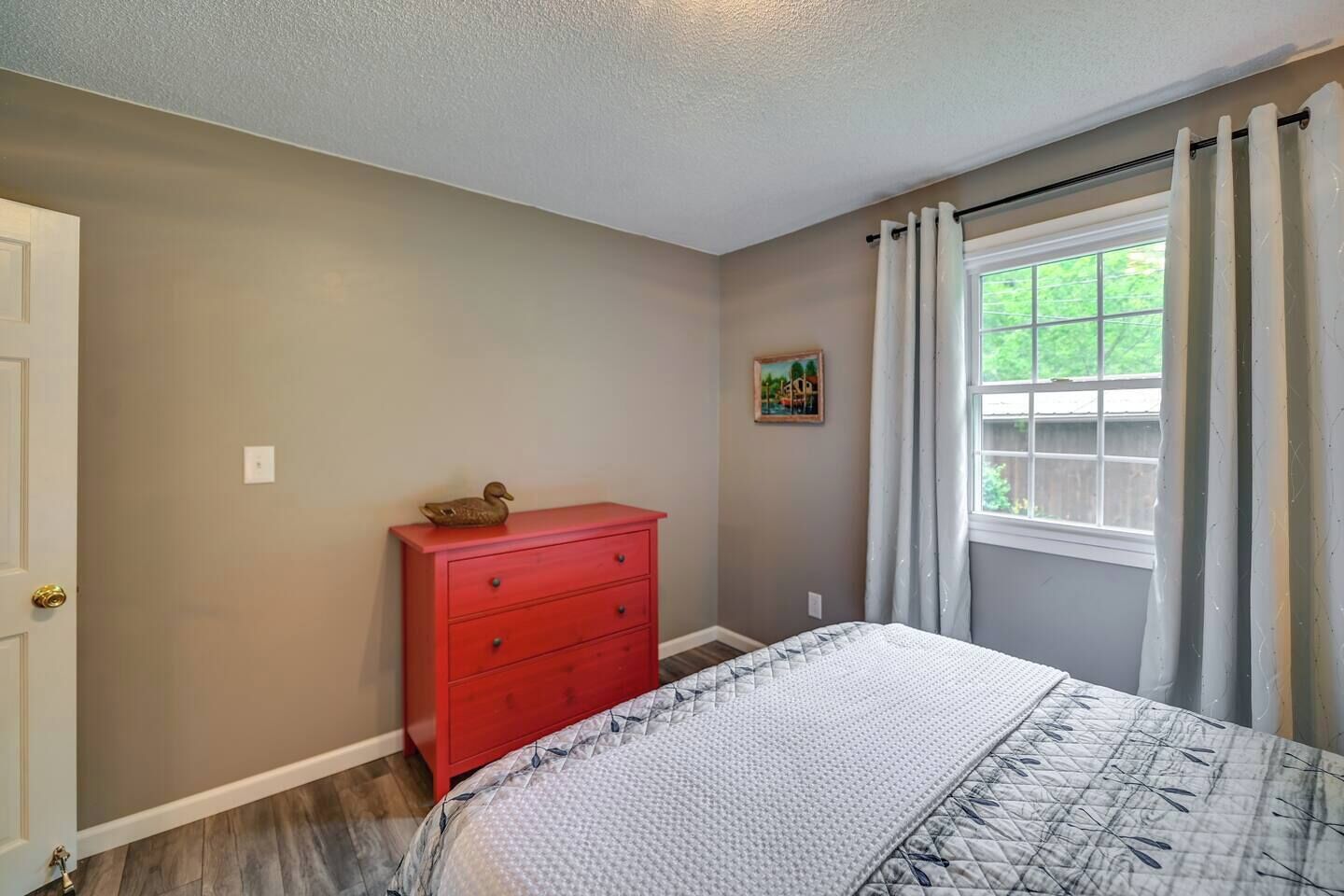 Un dormitorio con una cama, un aparador rojo, una ventana con cortinas y un cuadro pequeño en la pared.
