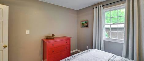 Un dormitorio con una cama, un aparador rojo, una ventana con cortinas y un cuadro pequeño en la pared.