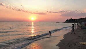 Escena de playa con el sol poniéndose sobre el océano, gente caminando por la orilla y una colina distante.