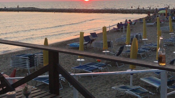Una playa con tumbonas y sombrillas, una terraza de madera y un atardecer sobre el océano.