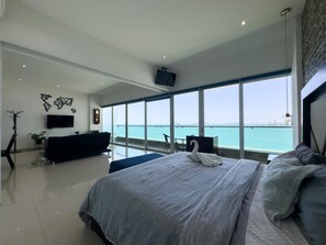 Un dormitorio moderno con una cama grande, un televisor, un balcón con vista al mar y un escritorio con una silla.