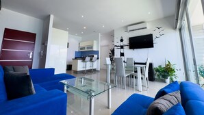 A modern living room with blue sofas, a glass coffee table, a dining area with white chairs, a kitchen with blue backsplash, and a TV mounted on the wall. - Beautiful Malecon and Beyond 514 Torre eMe (Mazatlán)