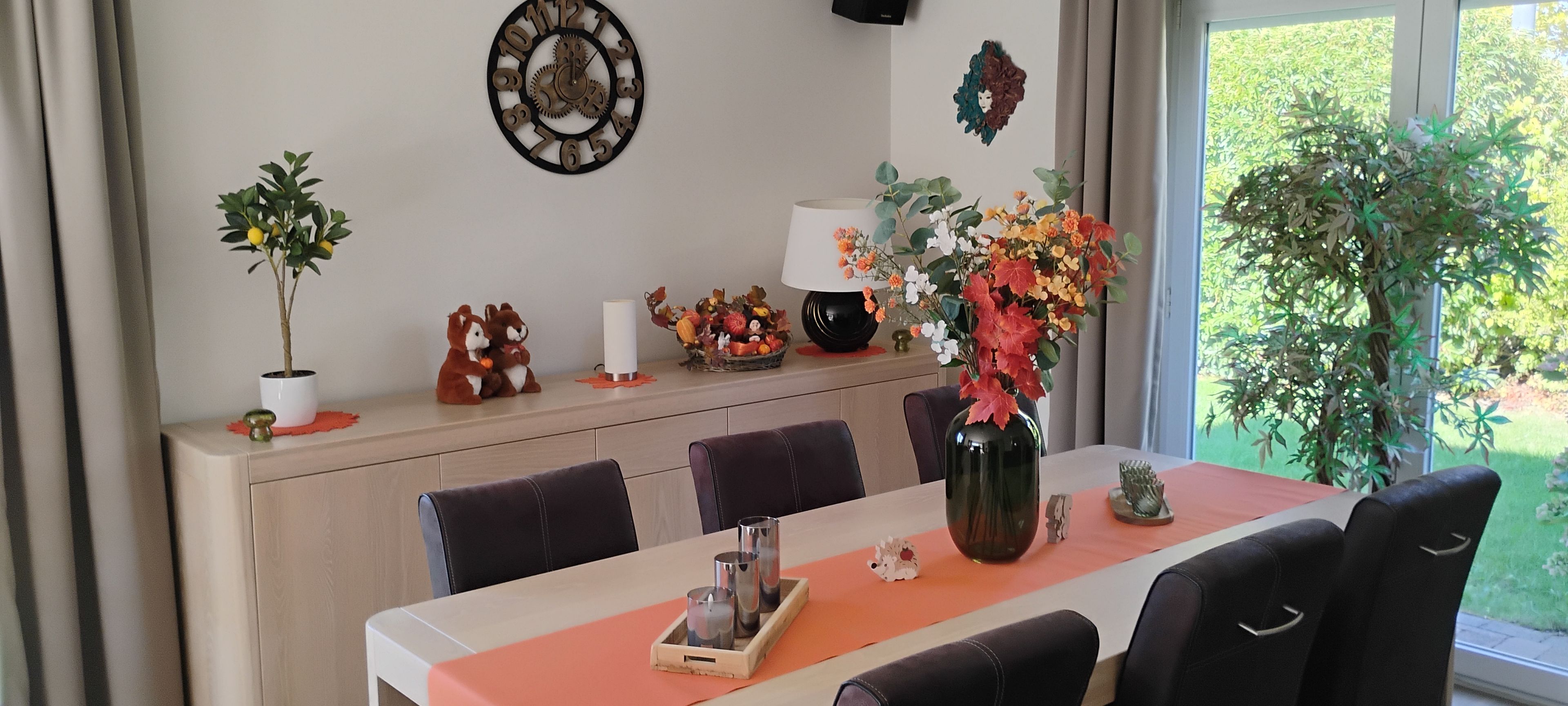 A dining area with a table set for four, a decorative wall clock, and a window with a view of greenery.