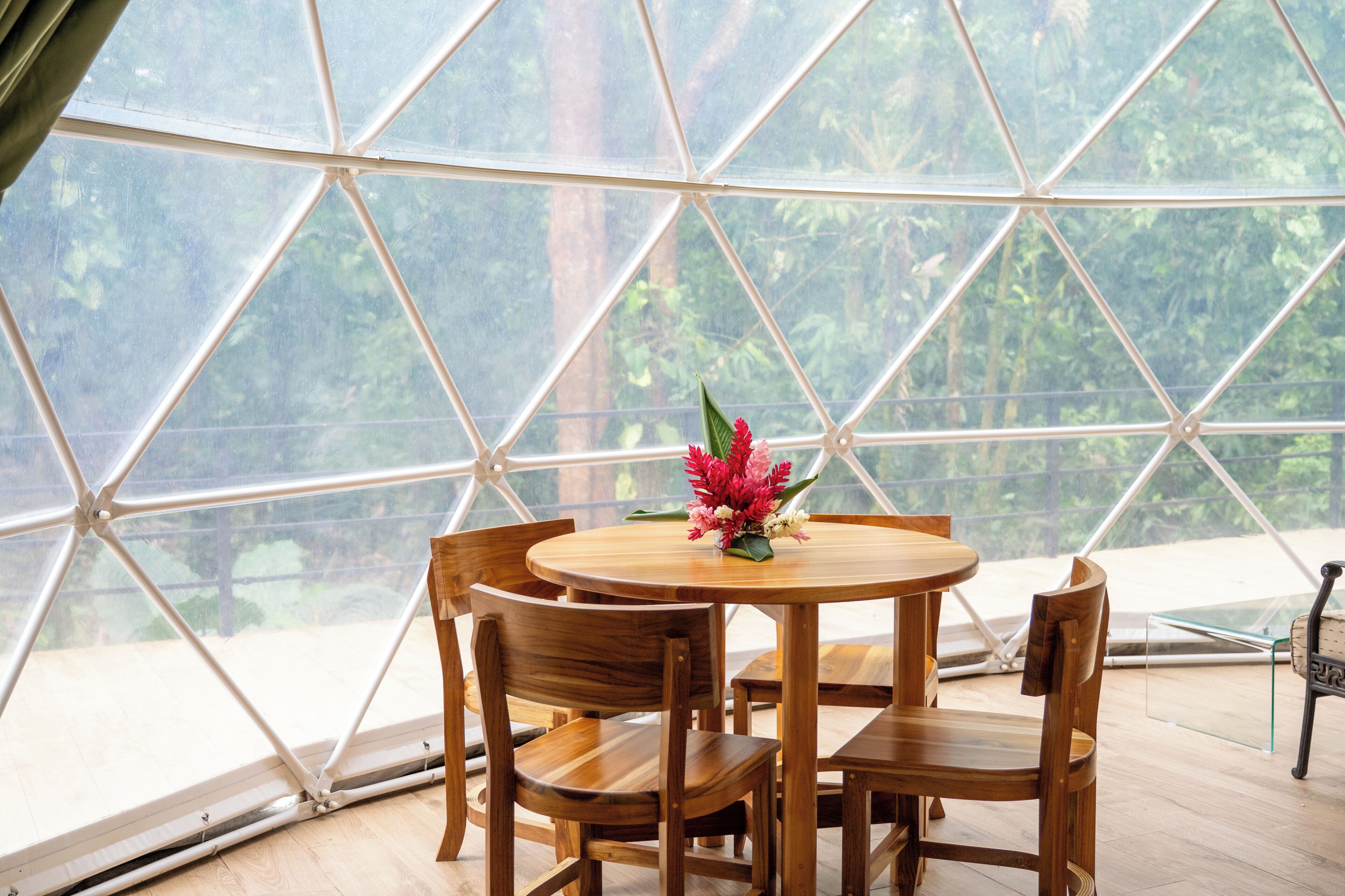 A round wooden table with chairs inside a geodesic dome structure, with a view of greenery outside.