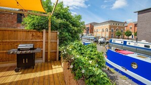 Une terrasse en bois avec un barbecue, un auvent jaune et une vue sur un canal où naviguent des bateaux.