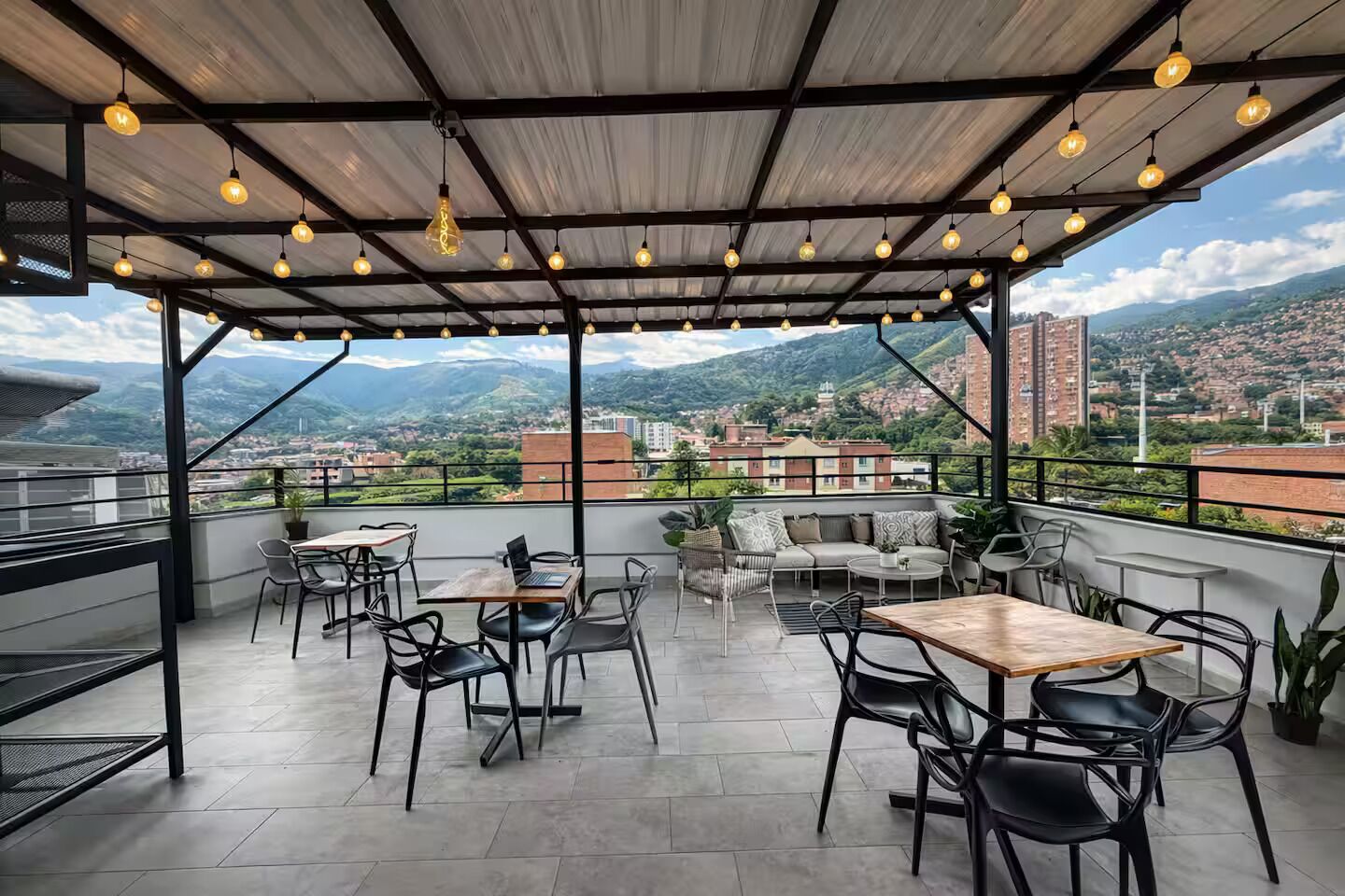 A rooftop terrace with tables and chairs, a view of a cityscape, and a metal roof structure.
