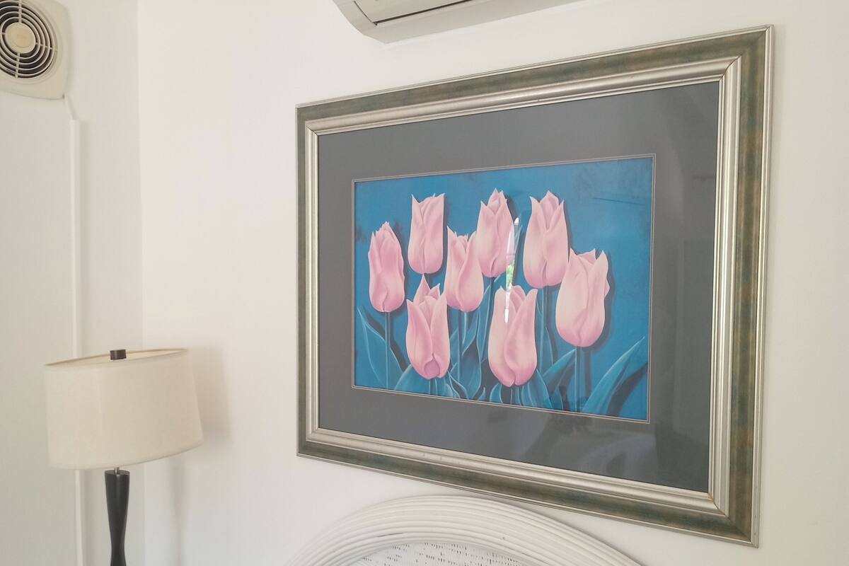 Quarto com cama, ar-condicionado fixado na parede, quadro com tulipas rosa e abajur.