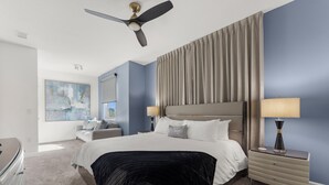 Une chambre d’hôtel avec un grand lit, un ventilateur de plafond, une table de chevet avec une lampe, une commode et un tableau au mur.