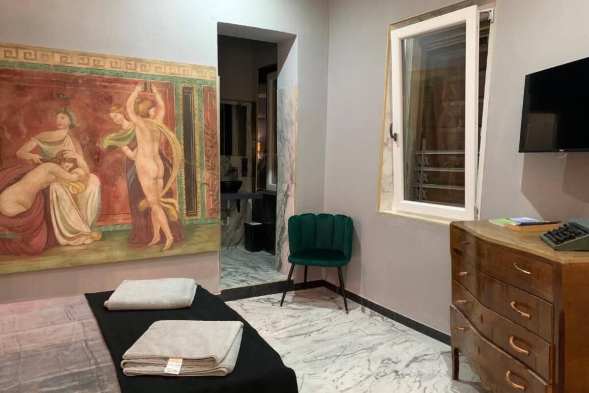 A room with a bed, a green chair, a wooden dresser, a painting, and a window.