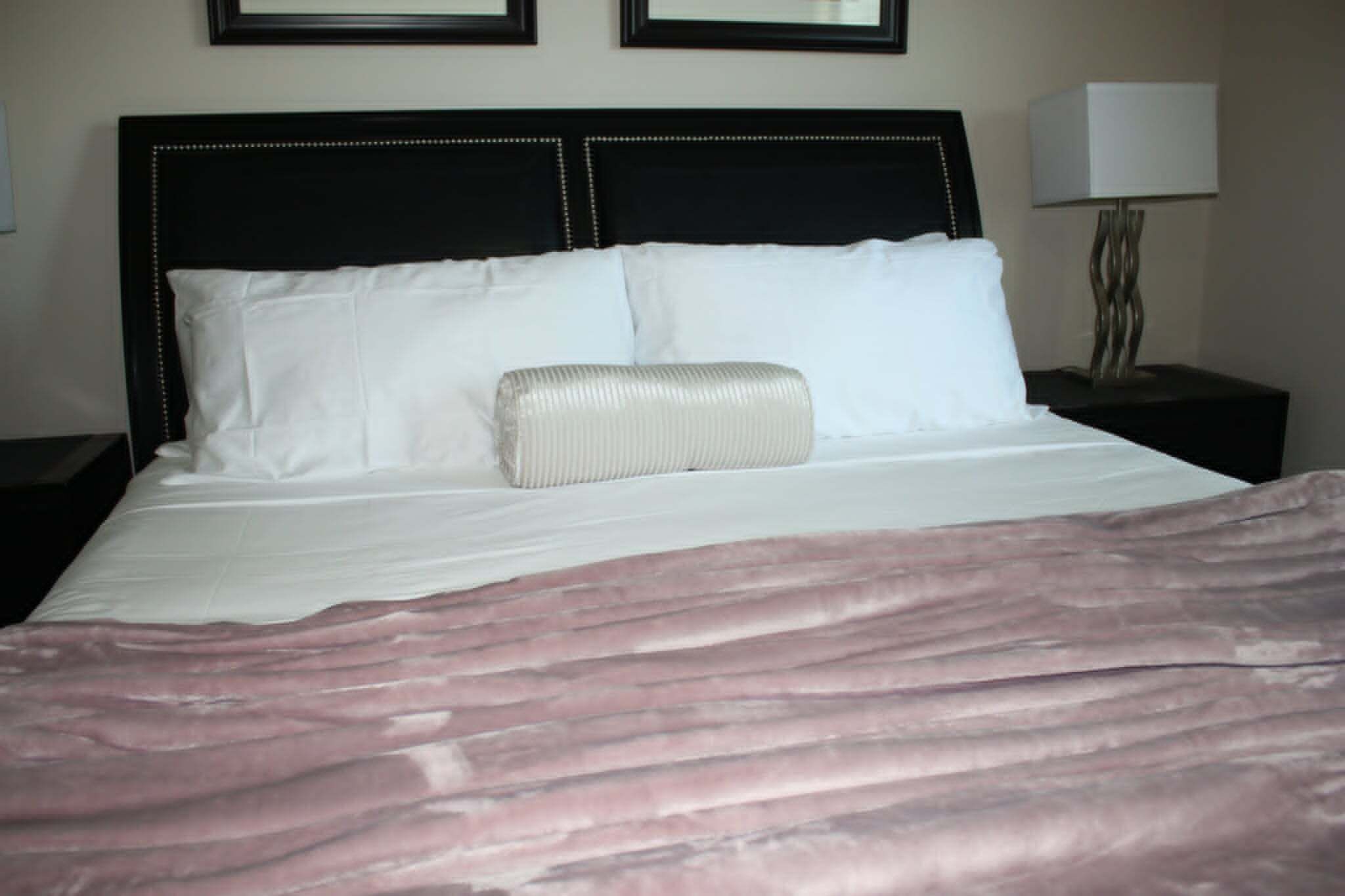 Una cama bien hecha con sábanas blancas y una manta rosa, un cabecero negro con tachuelas decorativas y una lámpara de noche en una mesita.