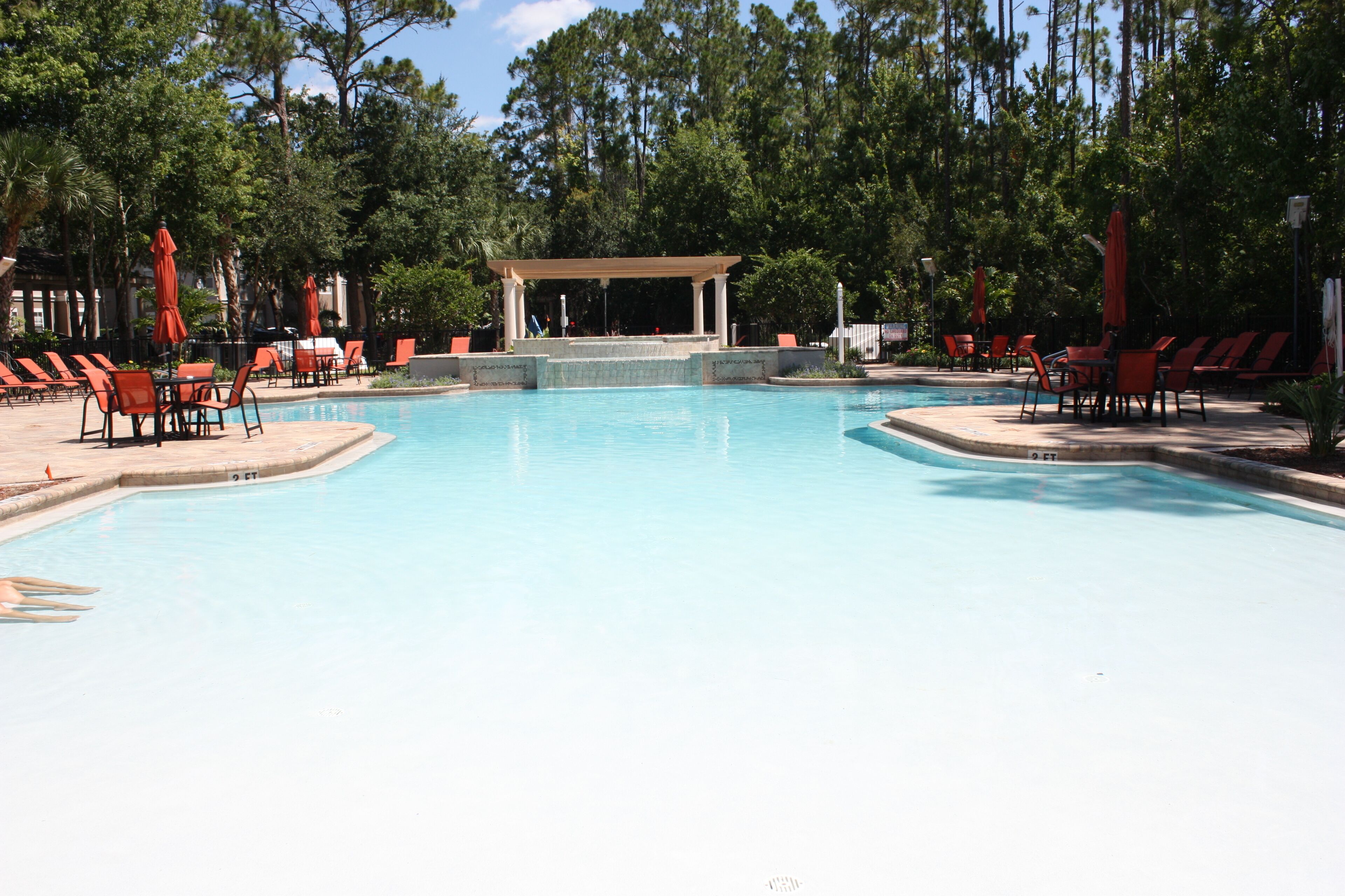Piscina con sillas de descanso y sombrillas, rodeada de árboles.