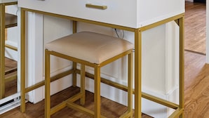 Un tocador blanco con marco dorado, un espejo redondo y una silla color beige.