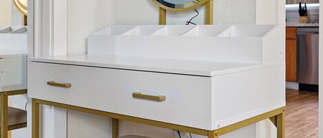 Une coiffeuse blanche avec un cadre doré, un miroir rond et une chaise beige.