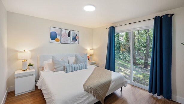 Un dormitorio con cama, mesitas de noche, lámpara, ventana con cortinas y vista a árboles.