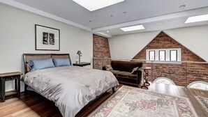 Um quarto com cama, mesa de cabeceira, cômoda, sofá, tapete estampado e parede de tijolos.