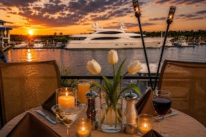A table set for a meal with candles, a vase with flowers, and glasses of wine, overlooking a marina with boats and a sunset. - Couples King Suite at Barefoot Resort / Private Entrance, Resort Pool, Marina (N Myrtle Beach)