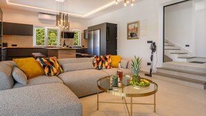 A modern living room with a sectional sofa, a glass coffee table, and a kitchen area with large windows.