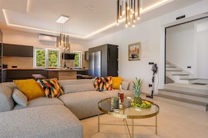 Ein modernes Wohnzimmer mit Ecksofa, Glastisch und Küchenzeile mit großen Fenstern.